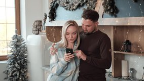 Happy young couple in love hugging in the kitchen decorated bright twinkle light and glowing xmas tree. Woman holds smartphone choosing gift on Christmas sale. Celebration atmosphere. Online Shopping. - Powered by Shutterstock - Get 15% off with code: PIKWIZARD15