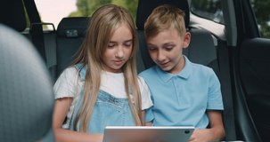 Portrait of nice friendly positive teen boy and girl which traveling by car on rear seat and browsing interesting video or movie on tablet pc
