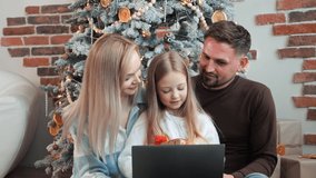 A young family of three people, father, mother and daughter are sitting near the Christmas tree making a video call using a laptop, listening her kid, little girl telling about her dream. Family time.