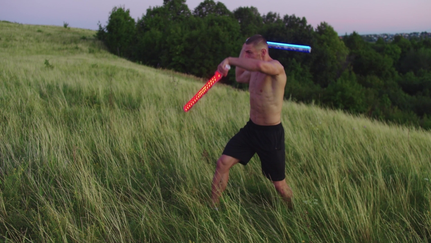 A muscular man in sunglasses waves two neon lightsticks like a light swords on a field