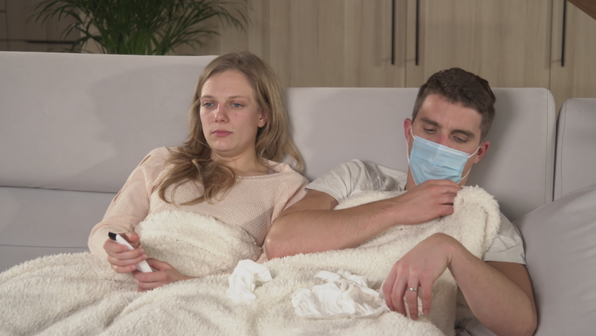 CLOSE UP: Couple on comfy sofa watching TV while guy is coughing and being sick. Young man wears protective mask and sneezes then lady disinfects the air with spray. Winter colds and flu spread around Royalty-Free Stock Footage #1096005867