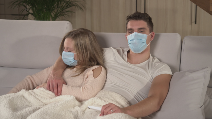 CLOSE UP: Lovely couple resting on comfy sofa and getting over a seasonal cold. Winter colds and flu spreading around. Young guy and lady being on sick leave, covered with blanket and watching TV. Royalty-Free Stock Footage #1096005883