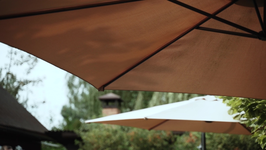 Large brown fabric canopy on spokes in the form of umbrella in the green courtyard of the house, close-up. The sun and rain protection dome sways in the wind. Royalty-Free Stock Footage #1096007339