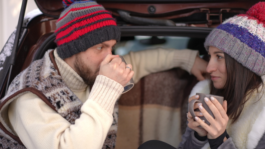 Side view portrait of happy young couple drinking hot tea sitting in car trunk on winter day. Positive relaxed Caucasian boyfriend and girlfriend talking enjoying coffee on travel outdoors