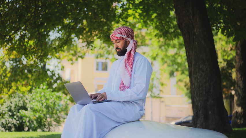 Serious Arab man in traditional middle-eastern clothes working on laptop in park