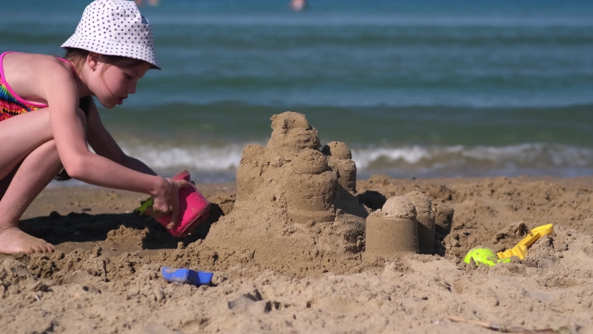 Little girl playing with sand on tropical beach. Kid at sea on summer family vacation. Sand and water fun, sun protection for children. Kid in swim suit build sand castle near sea or ocean Royalty-Free Stock Footage #1096023511