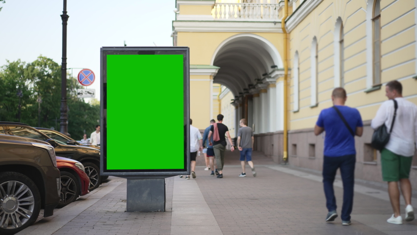 4k View of billboard and people walk on city street in summer spbd. Vertical digital display is standing by building, pedestrians walking and vehicles moving in modern town. Empty mockup poster