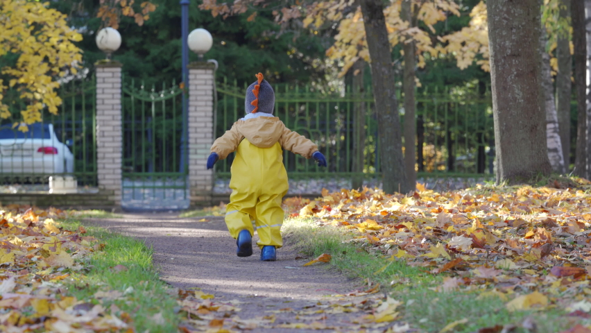 Happy toddler child in yellow rain pants running along the path in autumn park. Fall activities for toddlers.