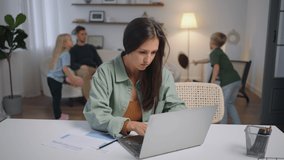 A business woman works remotely at home on a laptop while her family is singing in the background . Busy business woman. Father and children play
