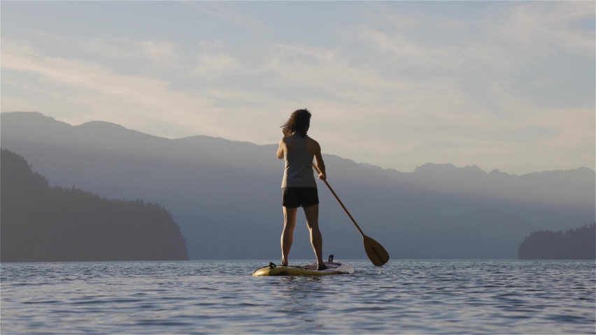 Adventurous Woman Paddling on a Paddle Board in a peaceful lake. Sunny Sunset. Harrison Hot Springs, British Columbia, Canada. Adventure Sport Travel Concept. Slow Motion Cinematic
