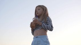 Trend video. A little girl in a vest drinks orange juice against the sky in the summer. Sunset sky. Girl Portrait View on sky in Summer Vacation. High quality FullHD footage