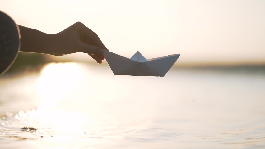 Woman hand holds paper boat on background of water on shore. child plays with paper boat near river, close-up.Launch boat on beach near the water. beautiful landscape of paper boat on water. Royalty-Free Stock Footage #1096037597