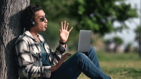 Smiling Spanish freelancer business man talking online video call in headphones laptop sitting on grass at summer park. Happy Arabic student guy e learning remotely classes teleworking communication