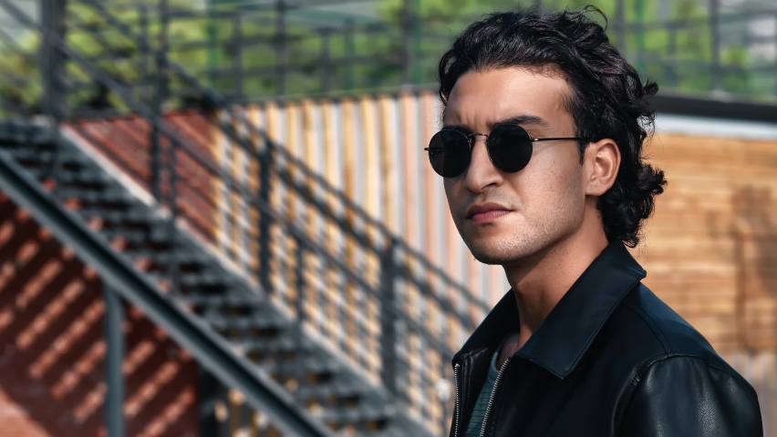 Brutal Latino curly man in sunglasses posing outdoor summer park wooden stairs construction closeup. Portrait confident handsome fashion guy in black leather jacket urban landscape sunny building