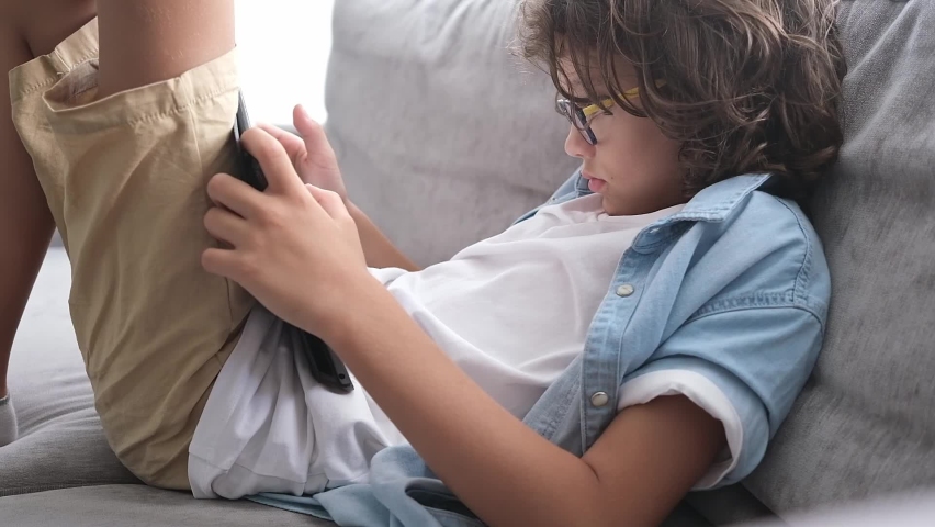 Little cute boy with eyeglasses and curly hair sitting on sofa and playing game on digital tablet. Portrait of child at home watching playing on the laptop. 