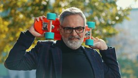 Grey-haired middle-aged white man wearing glasses and blazer standing at park holding red skateboard on his shoulder looking into camera smiling and showing thumbs up. Fun grandpa. Horizontal video