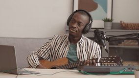 Medium shot of young male African American artist sitting at modern daw in home musical studio, signing in mic and playing guitar while having live stream