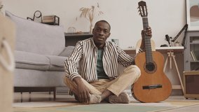 Lockdown of African American male musician sitting on floor at home with acoustic guitar in hand, looking at camera and speaking about guitar playing online