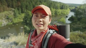 Chest up shot of Asian female hiker waving and speaking on camera while filming travel vlog in national park or web calling