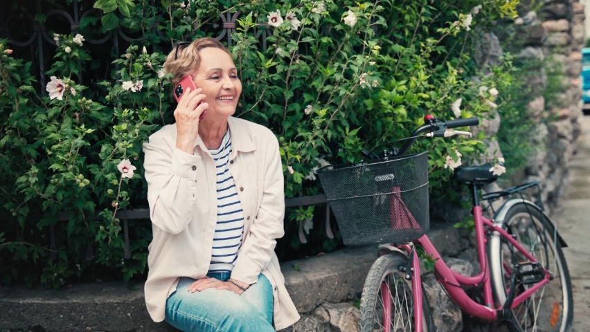 Portrait of a senior caucasian woman talking on her phone while sitting next to a bicycle on the side of the road in the city.