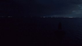 Bird's eye view of an old small sea lighthouse in the deep quiet calm Black Sea under the night cloudy dark sky near the sleepy town of Pomorie with bright lights in Bulgaria. UHD 4K video realtime