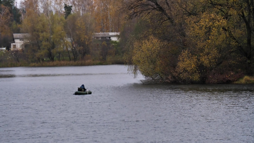 A fisherman on a boat in the middle of a lake against the background of yellowed trees and a village on a cloudy autumn day. Passion for fishing and outdoor activities. Industrial fishing.