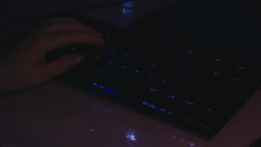 A woman in the dark is typing on a glowing computer keyboard. Working at a computer in a dark room. A woman's hand presses the keys on the keyboard.