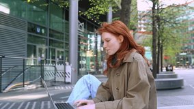 Happy teen redhead hipster girl student using laptop computer tech device on city street online learning studying outdoors, elearning outside watching educational webinar sitting in urban park.