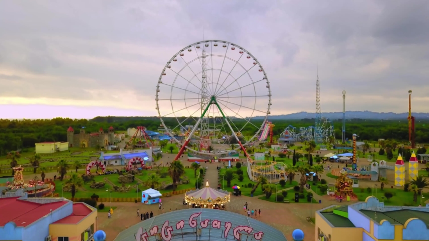 Top view of amusement park with Ferris wheel and roller coaster at sunset in Georgia at Tsitsinatela Park. Ferris wheel with air illumination. Recreation area of Georgian park on Black Sea coast. Royalty-Free Stock Footage #1096084323
