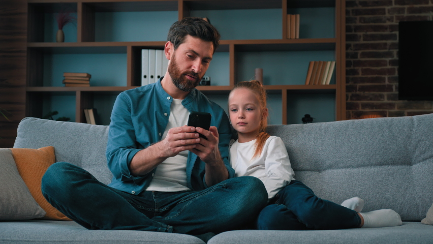 Caucasian dad and little daughter kid looking at mobile phone caring father teaching child girl using smartphone app play game online happy family rest together on sofa parental control tech education