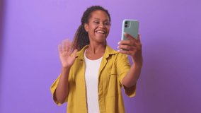 Young sociable cheerful African American woman student holding mobile phone and waving hand making video call to college or university friends discussing summer vacation, stands on purple background