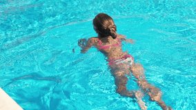 A beautiful funny slim girl in a bright pink swimsuit in children's purple glasses, floats in a large deep pool with clear transparent water on a warm sunny summer day