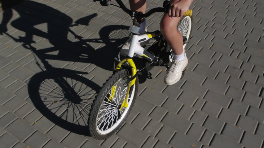 Girl rides a bicycle along the sidewalk on a sunny warm day. Beautiful shadow on the ground. Close-up of the hands on the steering wheel and the rotating front wheel. 200 fps slow motion Royalty-Free Stock Footage #1096092725