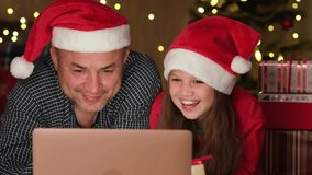 Happy family kid girl and father in Santa hat are laughing in front of laptop while online Christmas shopping or chatting in video chat. Daughter and dad watching funny movie and having fun at home