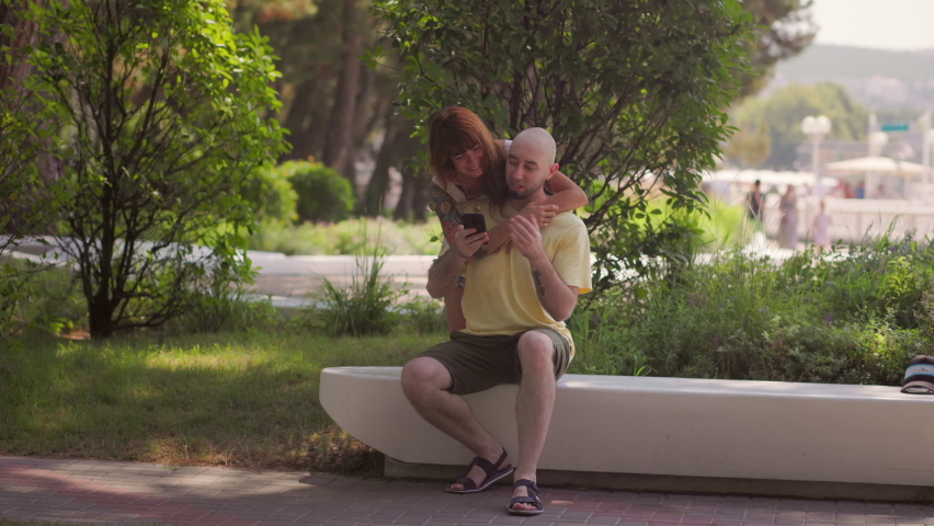 A couple of young Caucasian woman and man are sitting on a bench in the park, hugging and using the phone. Slow motion. The concept of relationship psychology.
