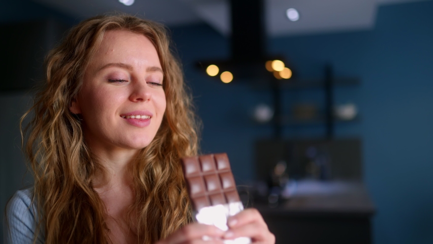 Hungry woman bites into a bar of chocolate and enjoys dessert. Curly blonde girl overeats unhealthy sweet food. Female stuffs her face into candy at kitchen interior. Healthy diet, overweight concept. Royalty-Free Stock Footage #1096105609