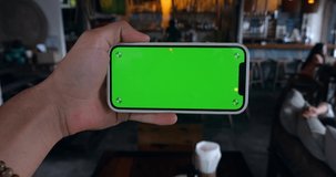 Man's hand holding smartphone in horizontal position with green screen chroma key indoors at cafe or restaurant background, close-up.