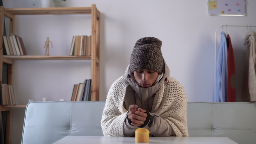 Tired man who warm hands over candlelight at home no heating. Shutdown of heating and electricity, power outage, blackout, load shedding or energy crisis.