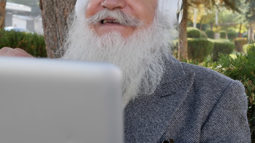 Cheerful grandfather with a gray fluffy beard and mustache is talking via video link using a laptop and white wireless headphones nodding and gesturing with his hands on a park bench at sunrise