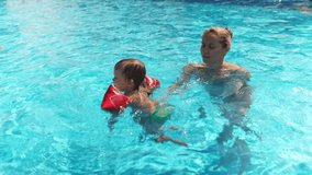 Happy caring kind mother in a bright bikini, plays and swims with a funny baby in oversleeves and his older sister in a deep blue pool with clear transparent water. HD slow-mo video
