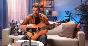 Cheerful Caucasian young talented man musician playing the guitar and recording video on smartphone while sitting on couch at home, video lesson online on cellphone in apartment, blogger concept