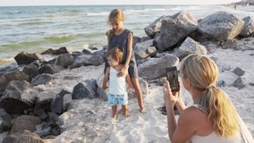 Beautiful happy kind mom takes pictures of her eldest fashionable caring daughter with her little cute cheerful brother on new modern touchscreen phone, on stone beach near sea. HD slow-mo video
