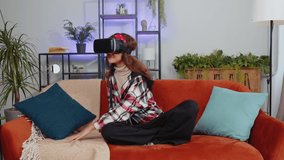 Preteen girl using virtual reality futuristic technology VR app headset helmet to play simulation 3D 360 video game, watching film movie at modern home apartment. Child kid in goggles sits on sofa