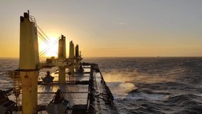 A silhouette video of a cargo ship out bulk carrier at sea during sunrise and during wind that make the sea spray blow 