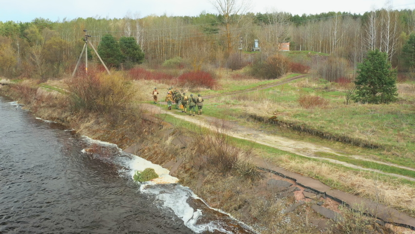 Men Dressed As US American Soldiers Of USA Infantry Of World War II Marching Walking Near River coast In spring Autumn Day. Group of Soldiers Marching In Forest. Aerial view elevated shot,