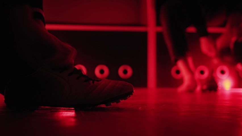 Soccer player puts football boots sneakers on his feet in dark locker room with red lighting. Cleats shoes close-up in dressing room. Amateur semi-professional leage. Cloakroom