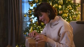 Beautiful happy brunette woman sits on armchair and opens gift box. Blurred Christmas tree with golden baubles in the background. Real time video. Selective focus. Winter holidays mood theme.