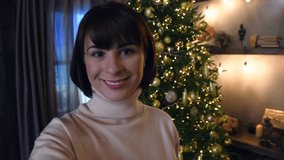 Selective focus. Beautiful caucasian brunette woman in white sweater records selfie video standing by Christmas tree at night. Real time video. People theme.
