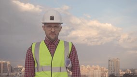 Attractive bearded male engineer or construction architect in helmet and safety reflective vest showing thumbs up gesture. Man holding blueprints in sunny day blue sky and urban city skyline. 4k video