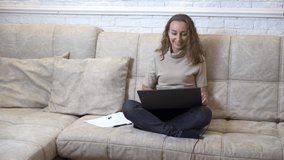 A young woman in a white cardigan sits on a sofa at home and looks at a laptop screen. The girl makes online purchases. Girl blogger communicates with her subscribers. The student calls the teacher
